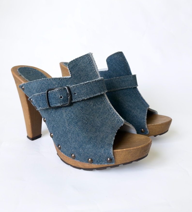 May include: A pair of denim clogs with wooden heels and soles. The blue denim uppers feature a buckle strap and open-toe design. The heels are approximately 4 inches tall, and the soles are studded with small metal rivets.