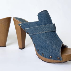 May include: A pair of denim clogs with wooden heels. The open-toe shoes feature a blue denim upper with a strap and are adorned with small, round metal studs. The heels are made of light brown wood.