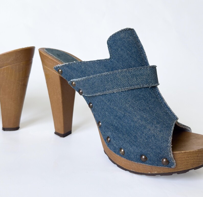 May include: A pair of denim clogs with wooden heels. The open-toe shoes feature a blue denim upper with a strap and are adorned with small, round metal studs. The heels are made of light brown wood.