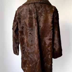 May include: A brown fur coat with a textured collar. The coat is a dark shade of brown and appears to be made of animal fur. The coat is displayed on a black stand against a white background.