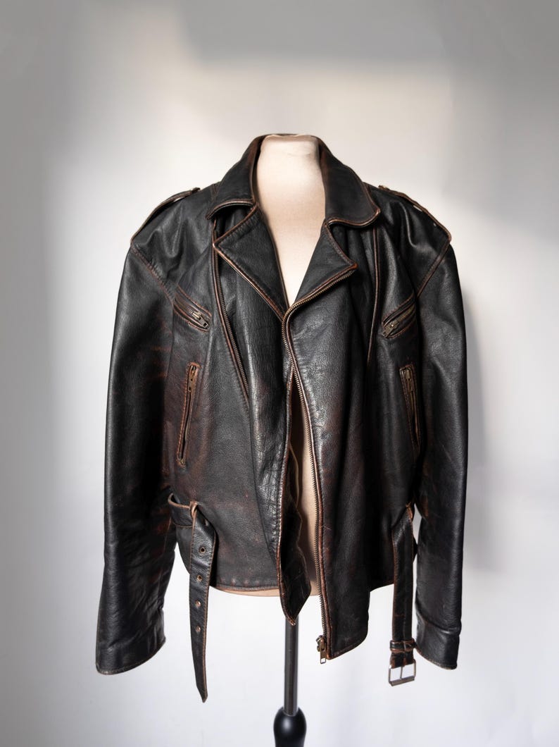 May include: A black leather motorcycle jacket with a classic asymmetrical zip closure. The jacket features multiple zippered pockets, shoulder epaulets, and a notched lapel. The leather has a distressed appearance with brown accents, and adjustable waist belts.