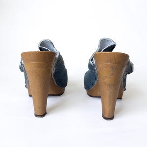 May include: A pair of denim clogs with wooden heels. The blue denim uppers feature a strap and rivet details. The wooden heels are a light brown color and have a slight flare. The clogs are set against a white background.