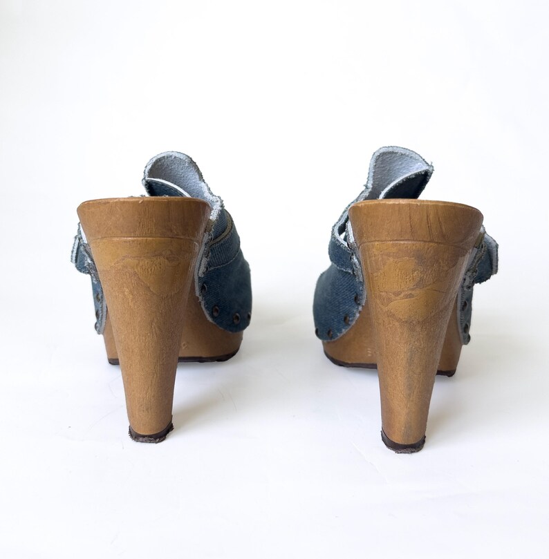May include: A pair of denim clogs with wooden heels. The blue denim uppers feature a strap and rivet details. The wooden heels are a light brown color and have a slight flare. The clogs are set against a white background.