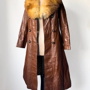 May include: A long, brown leather coat with a large, fluffy, tan and orange fur collar. The coat has a double-breasted design with dark buttons and a tailored fit. The coat is displayed on a mannequin.