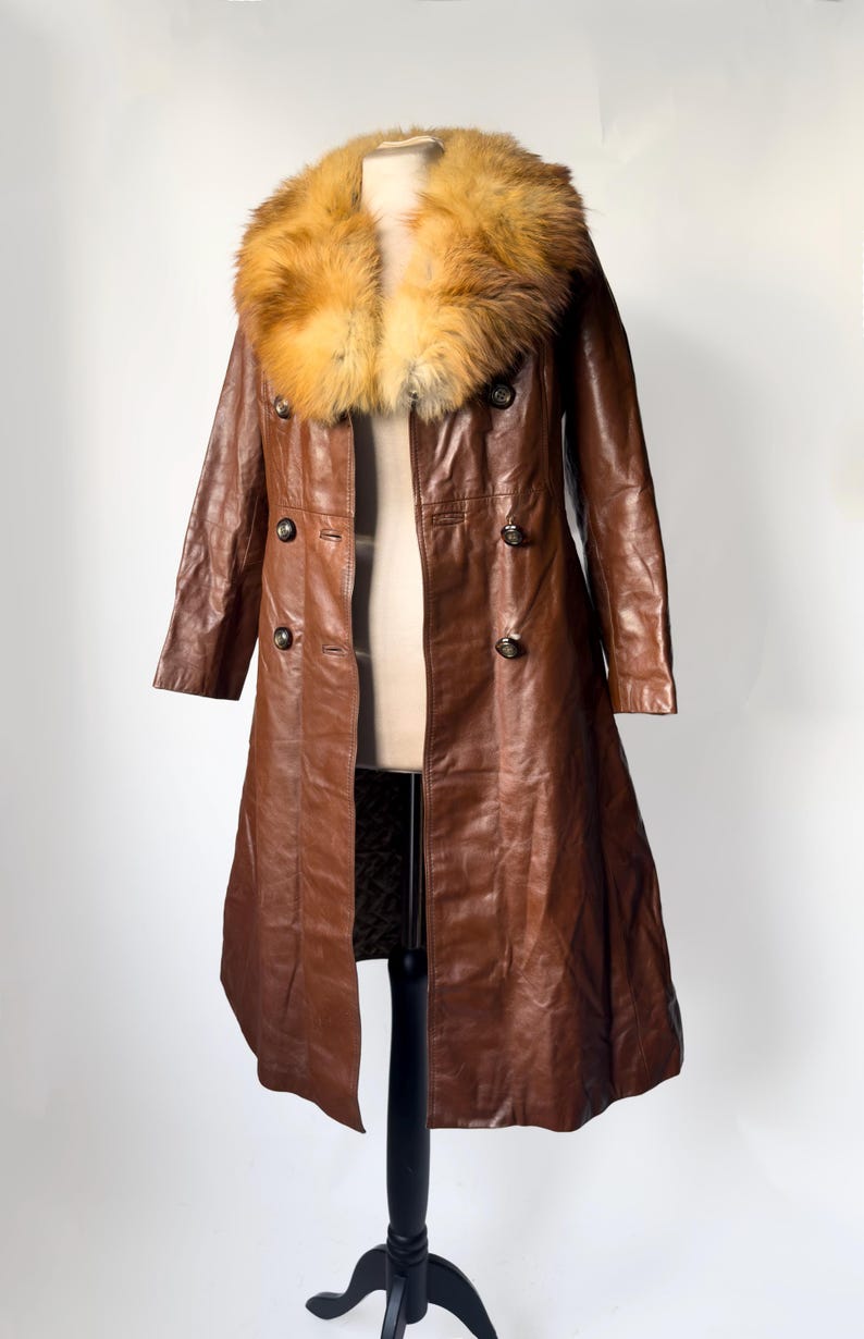 May include: A long, brown leather coat with a large, fluffy, tan and orange fur collar. The coat has a double-breasted design with dark buttons and a tailored fit. The coat is displayed on a mannequin.