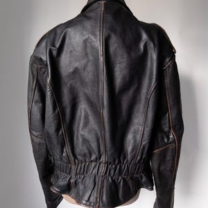 May include: A black leather jacket with a classic design, viewed from the back. The jacket features a collar, shoulder accents, and a gathered waist. Brown stitching and trim add contrast to the dark leather.