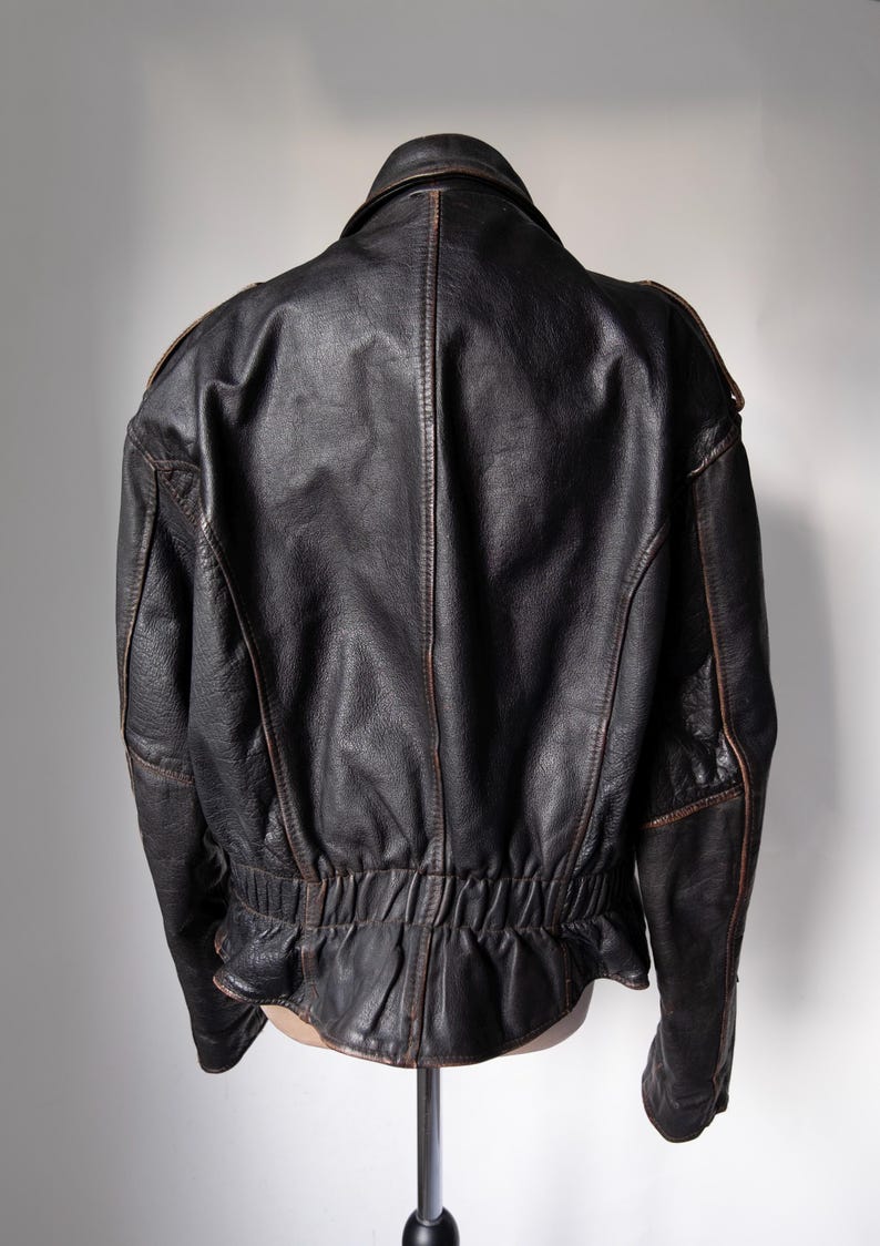 May include: A black leather jacket with a classic design, viewed from the back. The jacket features a collar, shoulder accents, and a gathered waist. Brown stitching and trim add contrast to the dark leather.