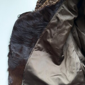 May include: A brown fur coat with a textured, curly collar. The coat's interior is a smooth, satin-like material. The fur appears to be a dark brown color, with a zipper detail visible on the side.