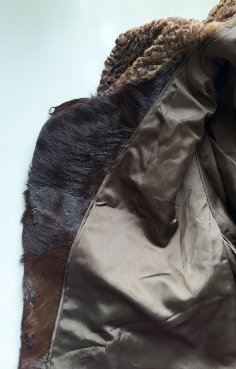 May include: A brown fur coat with a textured, curly collar. The coat's interior is a smooth, satin-like material. The fur appears to be a dark brown color, with a zipper detail visible on the side.