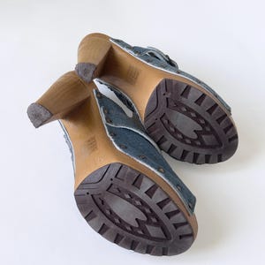 May include: A pair of denim clogs with wooden heels. The open-toe shoes feature a blue denim upper with metal stud accents and a brown rubber sole. The heels are made of light brown wood.
