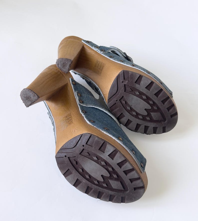 May include: A pair of denim clogs with wooden heels. The open-toe shoes feature a blue denim upper with metal stud accents and a brown rubber sole. The heels are made of light brown wood.