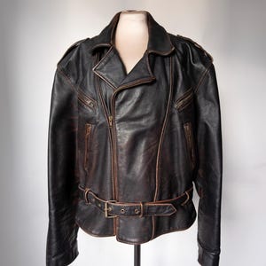 May include: A black leather jacket with a brown distressed finish. The jacket features a notched collar, an asymmetrical zipper closure, and multiple zippered pockets. It has a belt at the waist with a metal buckle. The jacket is displayed on a mannequin.