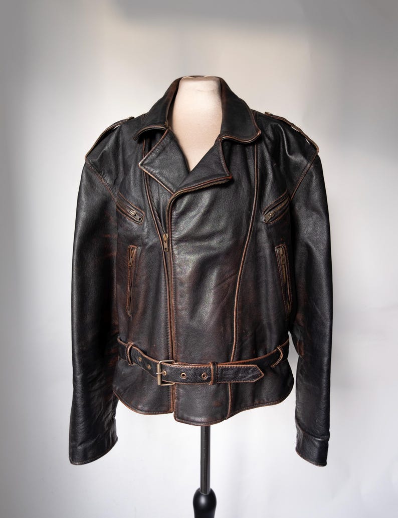 May include: A black leather jacket with a brown distressed finish. The jacket features a notched collar, an asymmetrical zipper closure, and multiple zippered pockets. It has a belt at the waist with a metal buckle. The jacket is displayed on a mannequin.