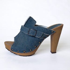 May include: Denim platform clogs with a wooden heel and sole. The open-toe shoe features a blue denim upper with a buckle strap and is adorned with small, round studs. The heel is approximately 4 inches (10 cm) high.