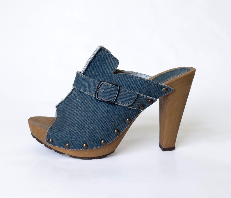 May include: Denim platform clogs with a wooden heel and sole. The open-toe shoe features a blue denim upper with a buckle strap and is adorned with small, round studs. The heel is approximately 4 inches (10 cm) high.