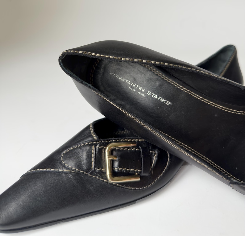 May include: Black leather flats with a gold buckle and a pointed toe. The shoes are by the brand Konstantin Starke, New York.