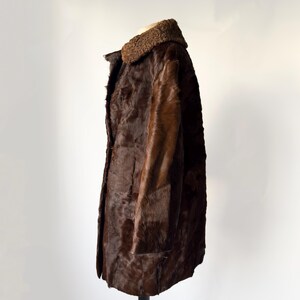 May include: A vintage brown fur coat with a textured appearance. The coat features a dark brown fur collar and a button-down front. The coat is displayed on a black stand against a white background.