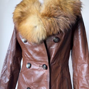 May include: A brown leather coat with a large, fluffy, light brown and tan fur collar. The coat has a double-breasted design with dark buttons and a tailored fit. The sleeves are long, and the coat appears to be vintage.