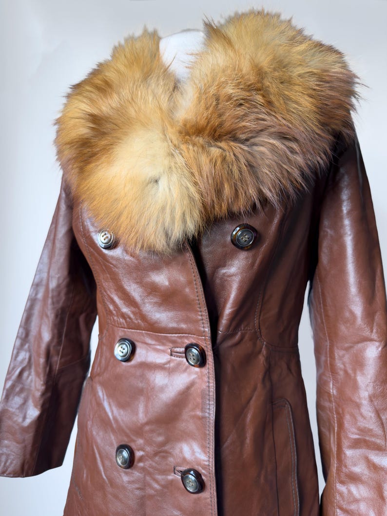 May include: A brown leather coat with a large, fluffy, light brown and tan fur collar. The coat has a double-breasted design with dark buttons and a tailored fit. The sleeves are long, and the coat appears to be vintage.