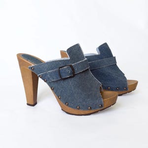 May include: A pair of denim clogs with wooden heels. The open-toe shoes have a buckle strap across the top and are adorned with small metal studs. The heels are approximately 4 inches high.