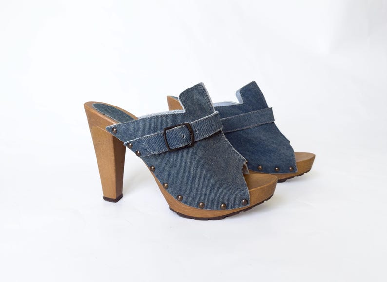May include: A pair of denim clogs with wooden heels. The open-toe shoes have a buckle strap across the top and are adorned with small metal studs. The heels are approximately 4 inches high.