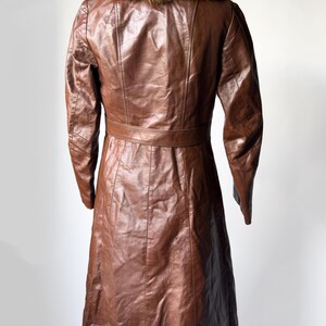 May include: A brown leather coat with a fur collar. The coat has a belted waist and a long, flowing skirt. The leather appears to be in good condition. The coat is displayed on a black stand against a white background.