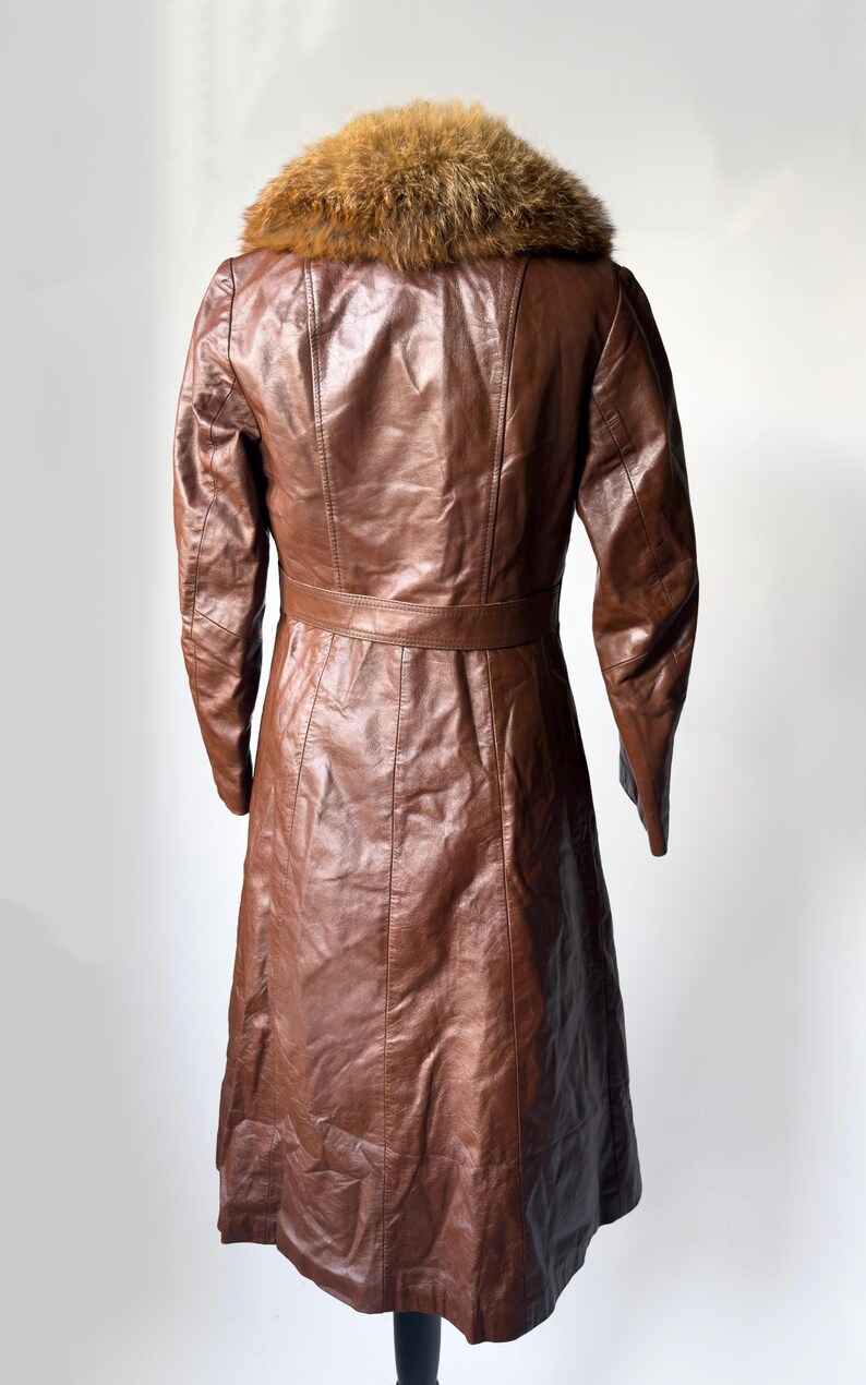 May include: A brown leather coat with a fur collar. The coat has a belted waist and a long, flowing skirt. The leather appears to be in good condition. The coat is displayed on a black stand against a white background.