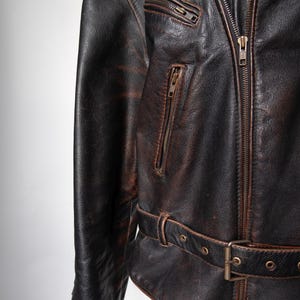 May include: Close-up of a distressed brown leather jacket. The jacket features a zipper closure, multiple zippered pockets, and a belt with a buckle at the waist. The leather has a worn, vintage appearance.