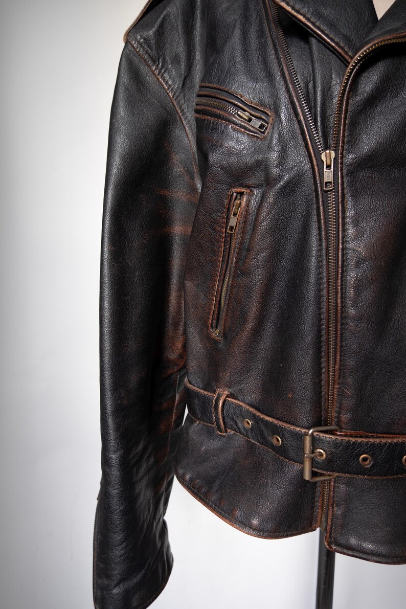 May include: Close-up of a distressed brown leather jacket. The jacket features a zipper closure, multiple zippered pockets, and a belt with a buckle at the waist. The leather has a worn, vintage appearance.