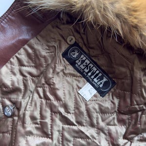 May include: Close-up of a brown leather jacket interior with a quilted lining. The jacket features a fur-trimmed hood and a label that reads "KESTILA MADE IN FINLAND". A button is visible on the jacket.
