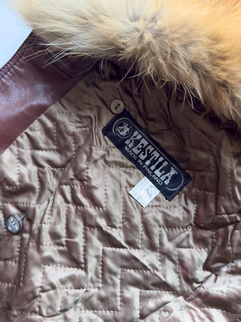 May include: Close-up of a brown leather jacket interior with a quilted lining. The jacket features a fur-trimmed hood and a label that reads "KESTILA MADE IN FINLAND". A button is visible on the jacket.