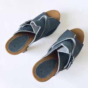 May include: A pair of denim sandals with wooden soles. The sandals feature a crisscross design across the top, with a light blue denim material and a natural wood-colored sole. The sandals are displayed on a white background.