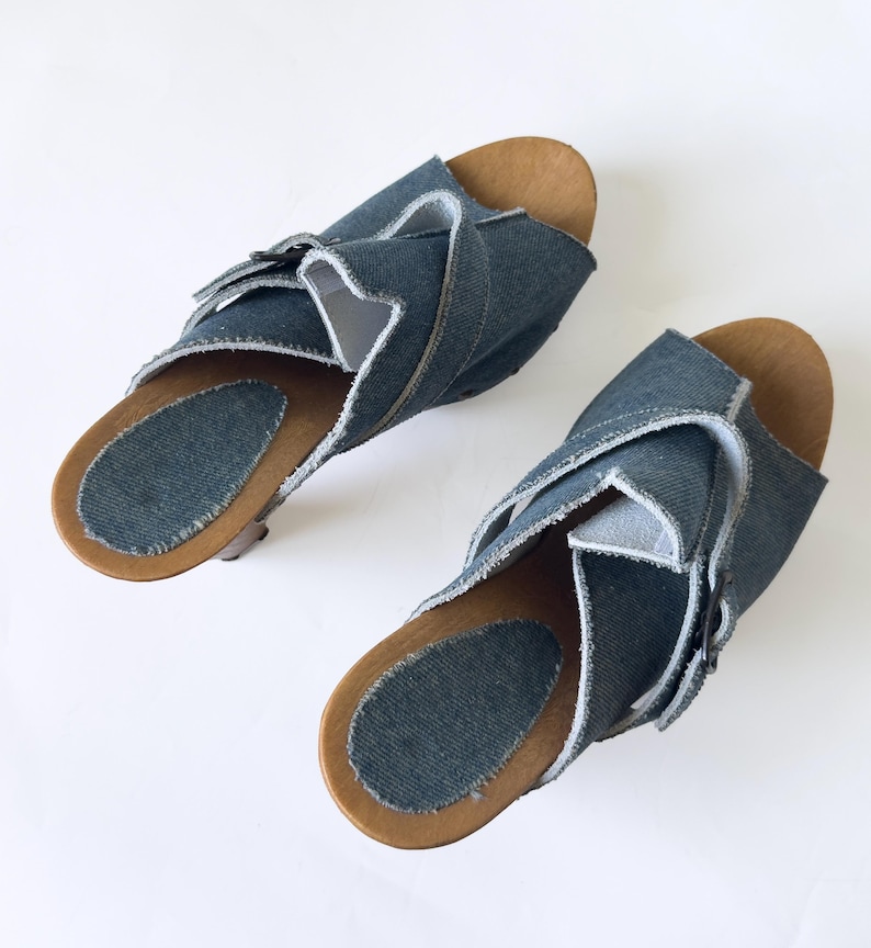 May include: A pair of denim sandals with wooden soles. The sandals feature a crisscross design across the top, with a light blue denim material and a natural wood-colored sole. The sandals are displayed on a white background.