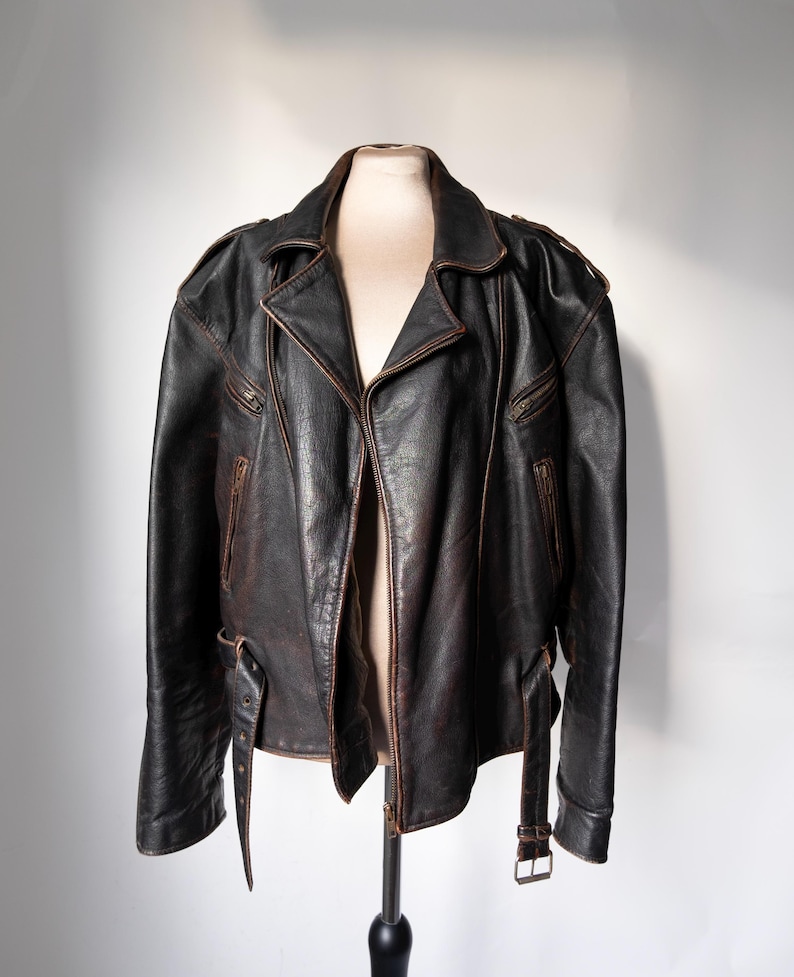 May include: A black leather jacket with a brown distressed finish. The jacket features a notched collar, asymmetrical zipper closure, and multiple zippered pockets. It has a belt at the waist and is displayed on a mannequin.