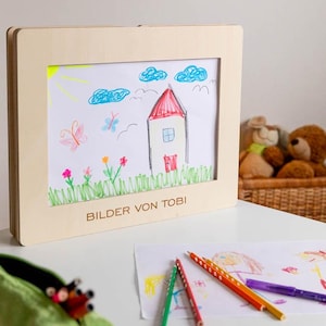 May include: A child's drawing of a house, clouds, and flowers in a wooden frame. The drawing is in bright colors, with the text "BILDER VON TOBI" at the bottom. Colored pencils and a green pencil case are on the table.