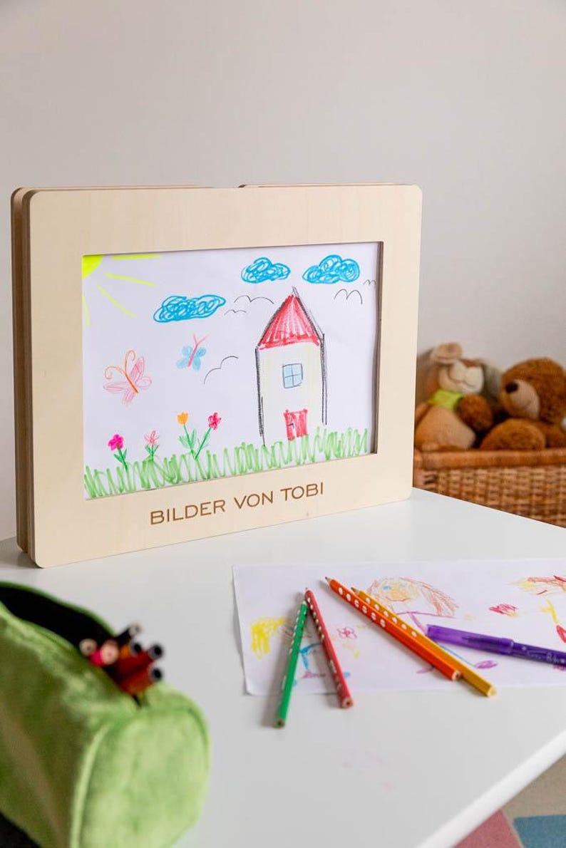 May include: A child's drawing of a house, clouds, and flowers in a wooden frame. The drawing is in bright colors, with the text "BILDER VON TOBI" at the bottom. Colored pencils and a green pencil case are on the table.
