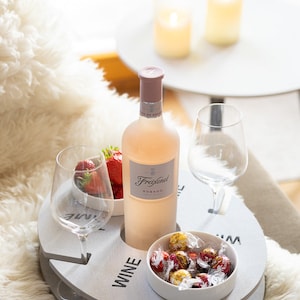 Könnte beinhalten: Ein weißes Holztablett mit einem integrierten Weinflaschenhalter und zwei Glashaltern. Das Tablett hat außerdem eine kleine Schale mit Schokoladenbonbons und eine Schale mit Erdbeeren. Eine Flasche Freixenet Rosado Wein steht auf dem Tablett.
