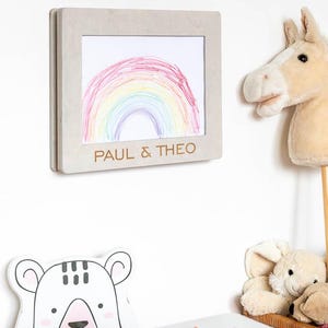 May include: A framed drawing of a rainbow in a child's room. The frame is light gray and has the names "PAUL & THEO" engraved on it. A stuffed horse head on a stick and a bear-shaped decoration are also visible.