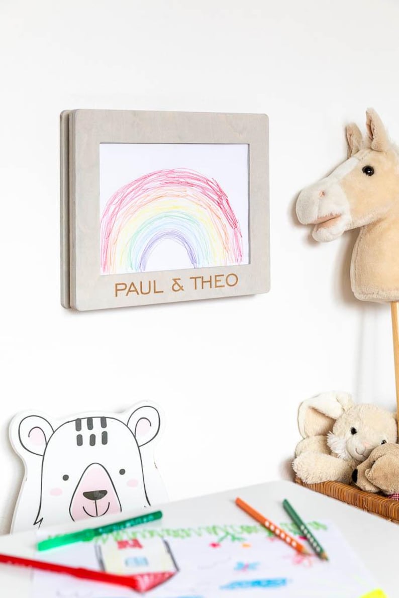 May include: A framed drawing of a rainbow in a child's room. The frame is light gray and has the names "PAUL & THEO" engraved on it. A stuffed horse head on a stick and a bear-shaped decoration are also visible.