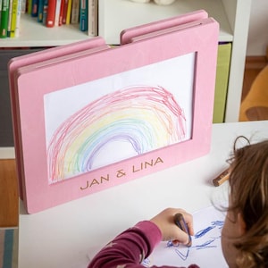 May include: A pink wooden frame displaying a child's colorful rainbow drawing. The frame has the text "JAN & LINA" engraved on it. A child is drawing with a crayon on a white surface.