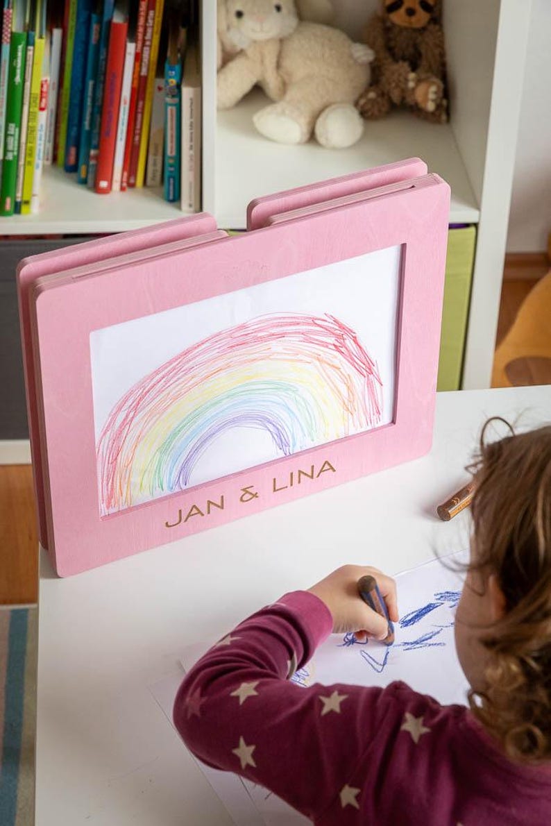 May include: A pink wooden frame displaying a child's colorful rainbow drawing. The frame has the text "JAN & LINA" engraved on it. A child is drawing with a crayon on a white surface.