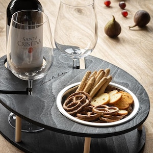 May include: A black wooden serving tray with two wine glass holders and a small bowl for snacks. The tray has a wooden base with two wooden legs. The tray is filled with snacks, including pretzels, breadsticks, and crackers. The tray is on a wooden table.
