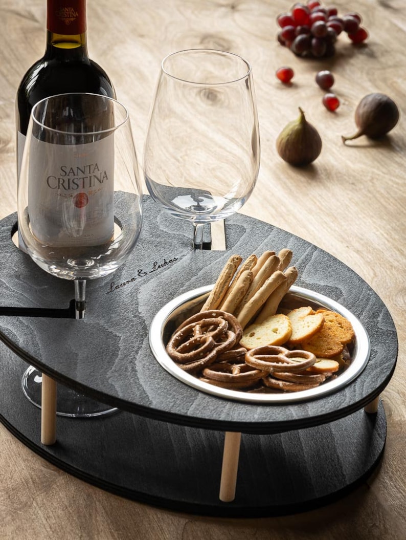 May include: A black wooden serving tray with two wine glass holders and a small bowl for snacks. The tray has a wooden base with two wooden legs. The tray is filled with snacks, including pretzels, breadsticks, and crackers. The tray is on a wooden table.