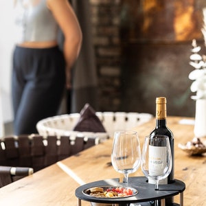 May include: A black wooden serving tray with two wine glasses, a bottle of wine, and a bowl of snacks. The tray has a built-in wine bottle holder and two slots for glasses. The tray is on a wooden table.