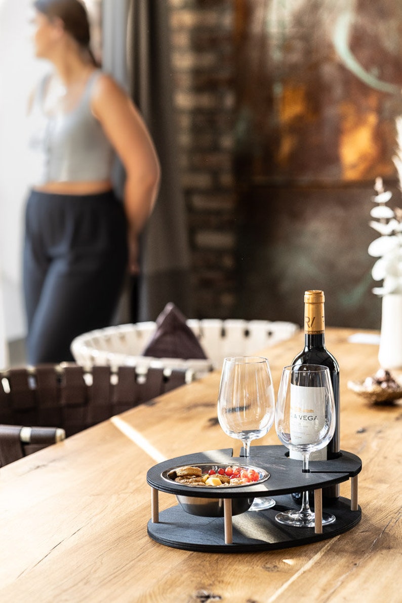 May include: A black wooden serving tray with two wine glasses, a bottle of wine, and a bowl of snacks. The tray has a built-in wine bottle holder and two slots for glasses. The tray is on a wooden table.