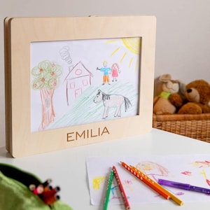 May include: A framed child's drawing featuring a house, tree, sun, and a horse. The frame is made of light-colored wood, with the name "EMILIA" engraved at the bottom. Colored pencils and other drawings are visible on the table.