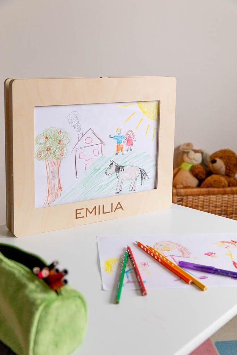 May include: A framed child's drawing featuring a house, tree, sun, and a horse. The frame is made of light-colored wood, with the name "EMILIA" engraved at the bottom. Colored pencils and other drawings are visible on the table.