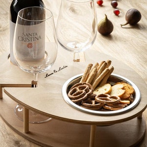 May include: A wooden serving tray with two wine glass holders, a small bowl for snacks, and the text "Luca & Lucia". The tray is on a wooden table with a bottle of wine, two wine glasses, grapes, and figs.