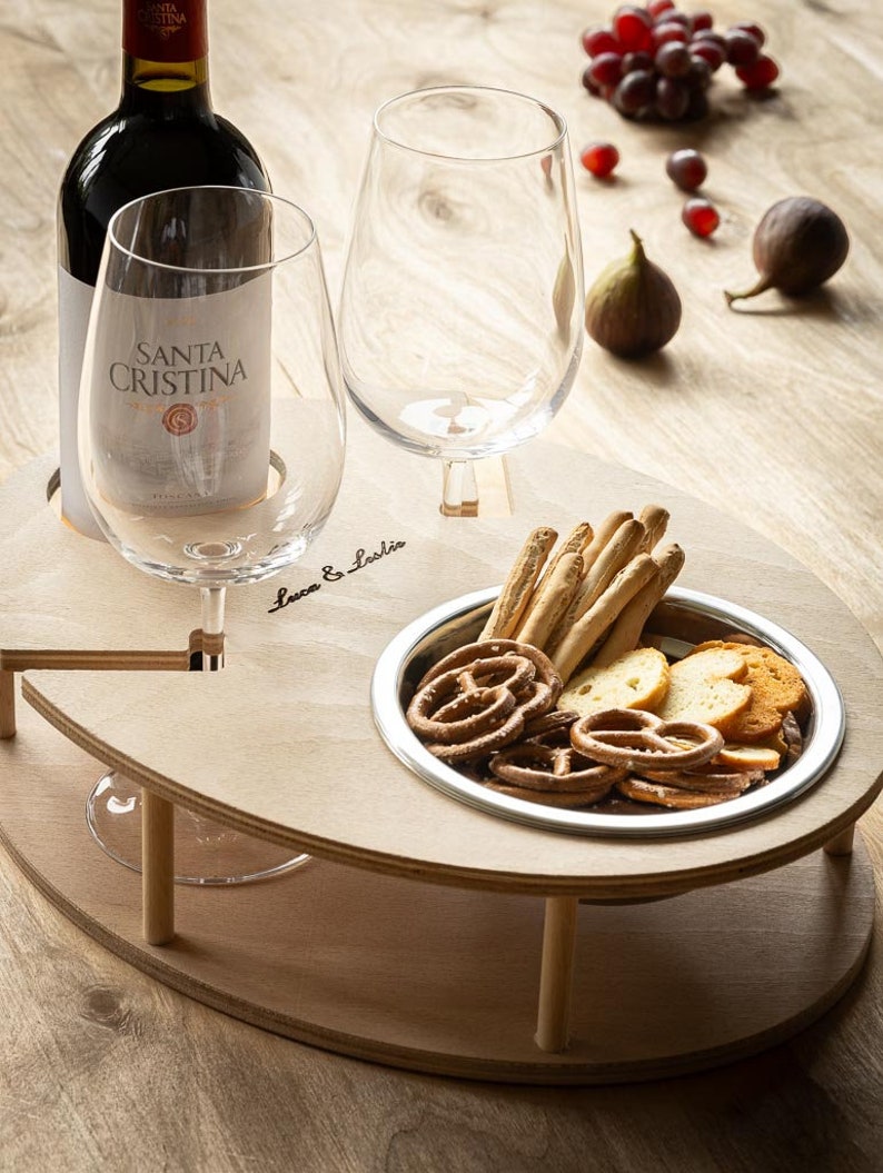 May include: A wooden serving tray with two wine glass holders, a small bowl for snacks, and the text "Luca & Lucia". The tray is on a wooden table with a bottle of wine, two wine glasses, grapes, and figs.