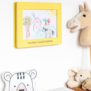 May include: A yellow framed drawing of a house, tree, people, and a horse, with the text "MAYAS KUNSTWERKE". A stuffed horse head and a bear-shaped decoration are also visible.